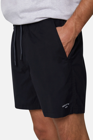 A person is wearing Industrie The Bahama Angler Shorts in black, featuring an elastic waist and "INDUSTRIE 1999" written in white on the lower left hem. Their right hand is in their pocket, and they are also wearing a light-colored shirt.
