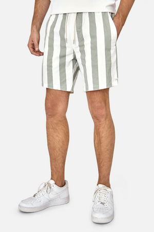 An individual dons Industrie The Beverly Bahama Short, featuring green and white vertical stripes with a drawstring and elastic waist. They wear white sneakers; the upper body is mostly cropped out, set against a plain light gray backdrop.