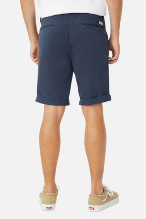 A person stands facing away, donning a relaxed fit white shirt with navy blue Industrie The Rinse Drifter Shorts made of cotton elastane, featuring rolled cuffs, paired with beige canvas sneakers with white soles. The background is plain white.