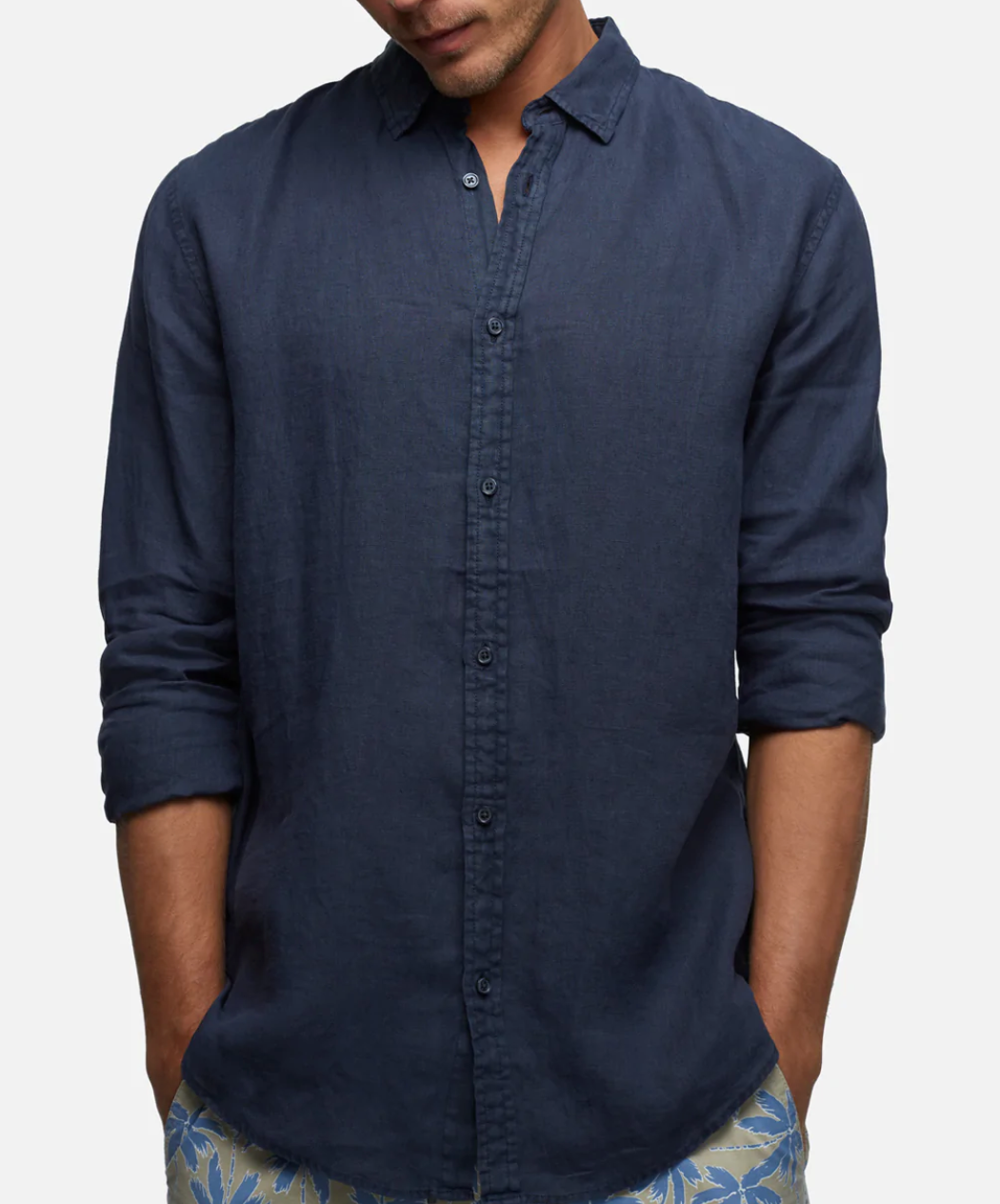 A person in a dark blue, relaxed fit Industrie The Tennyson Linen Shirt with rolled-up sleeves stands against a plain background. Their hands are in their pockets, and they are wearing patterned shorts. The face is not visible.