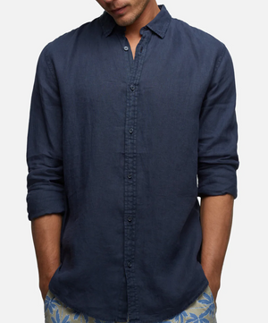 A person in a dark blue, relaxed fit Industrie The Tennyson Linen Shirt with rolled-up sleeves stands against a plain background. Their hands are in their pockets, and they are wearing patterned shorts. The face is not visible.