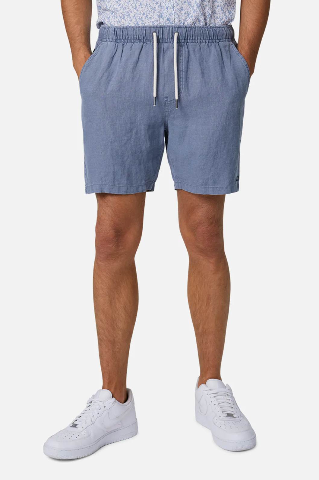 A person wearing Industrie The Marina Linen Short in light blue, featuring an elastic drawstring waist, stands with hands in pockets paired with white sneakers and a light top on a plain white background.