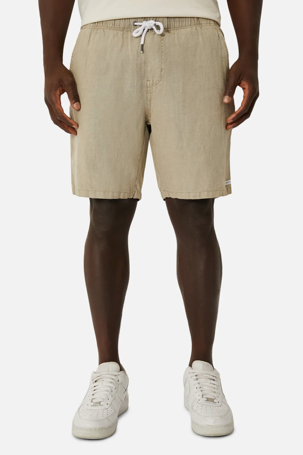The lower half of a person wearing Industrie The Marina Linen Shorts in light beige with an elastic waist and drawstring, paired with white sneakers, is visible against a plain white background.