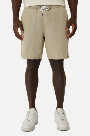 The lower half of a person wearing Industrie The Marina Linen Shorts in light beige with an elastic waist and drawstring, paired with white sneakers, is visible against a plain white background.