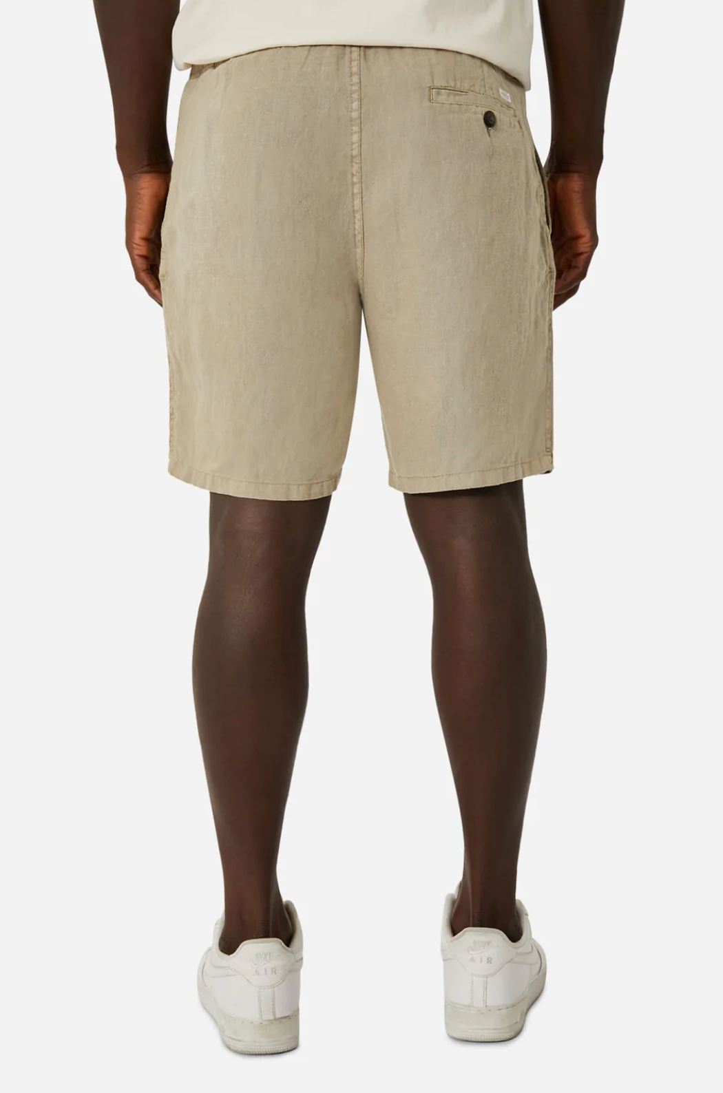 Rear view of a person wearing Industrie The Marina Linen Short in beige with an elastic waist and visible back pockets. They pair it with white sneakers and a tucked-in white shirt, all set against a white background.