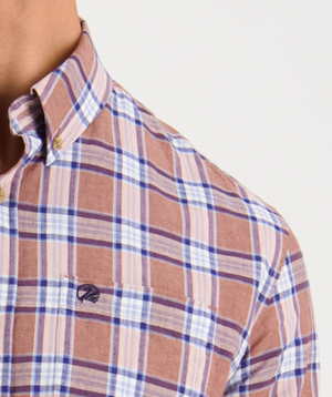 A person in a Swanndri Kennedy Point Shirt with a tailored fit, showcasing a brown, blue, and white plaid pattern. It features an embroidered chest logo and is made from a comfortable cotton-linen blend. The top button is open for casual style against a plain white background.
