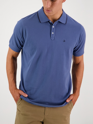 A man in a Swanndri Pinehurst Polo, a blue 100% cotton shirt with a black trim collar and embroidered logo, easily pairs with khaki pants. He looks down, hands in pockets against a plain white background.