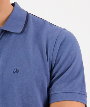 The image shows a close-up of a person wearing the Swanndri Pinehurst Polo, a blue 100% cotton shirt with an embroidered Swanndri logo on the chest and dark trim on the collar. The focus is on the upper torso, including part of the left shoulder.