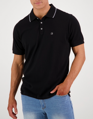 A person in a Swanndri Pinehurst Polo, made of black 100% cotton with dark buttons and a small embroidered logo on the chest, stands with one hand in their blue jeans pocket against a plain white background.