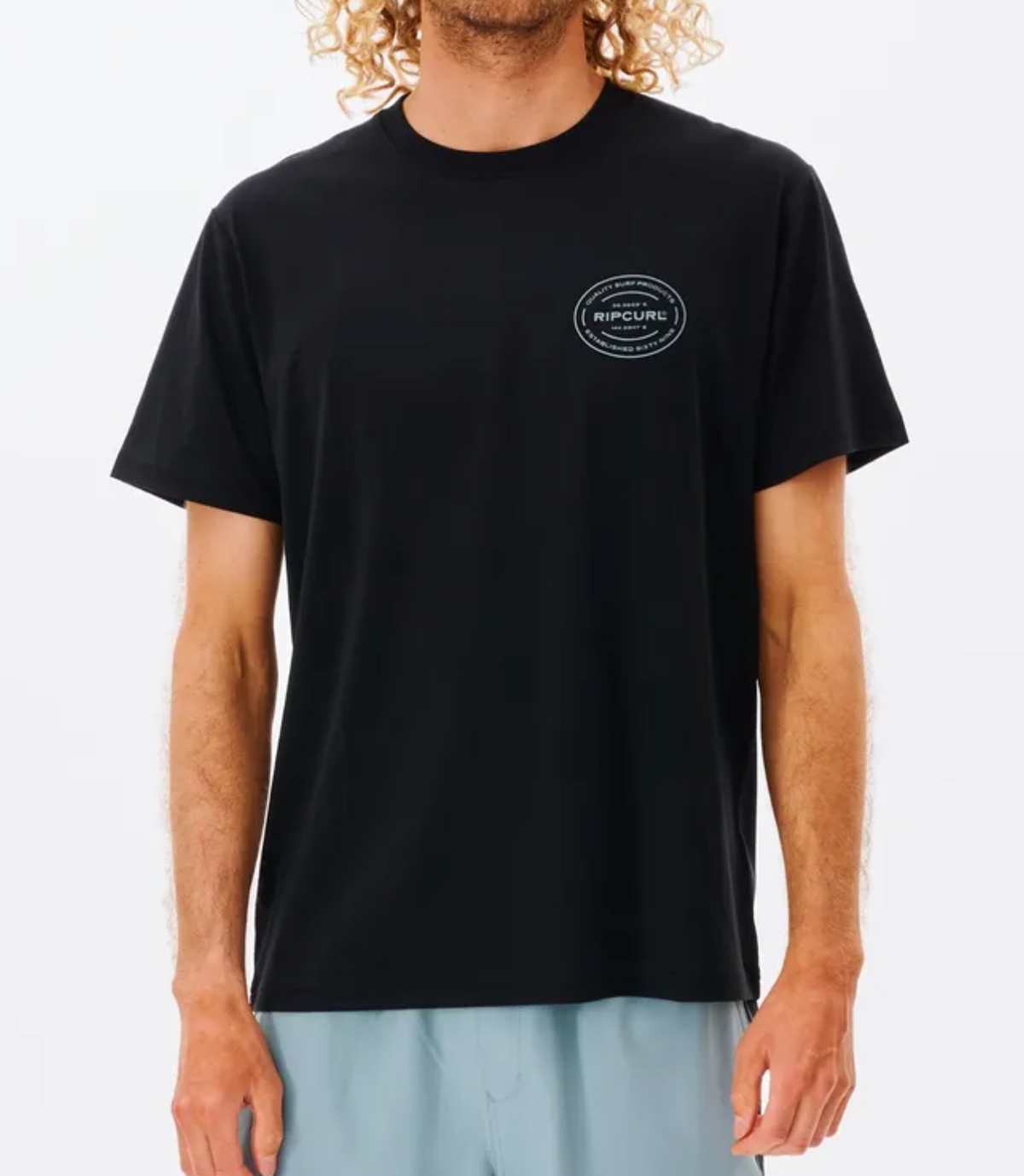 A person with curly hair wears the black Rip Curl Vaporcool Oval Tee, featuring a small circular chest logo and light blue shorts. Its quick-dry, moisture-wicking fabric is ideal for an active lifestyle, shown against a plain white background.
