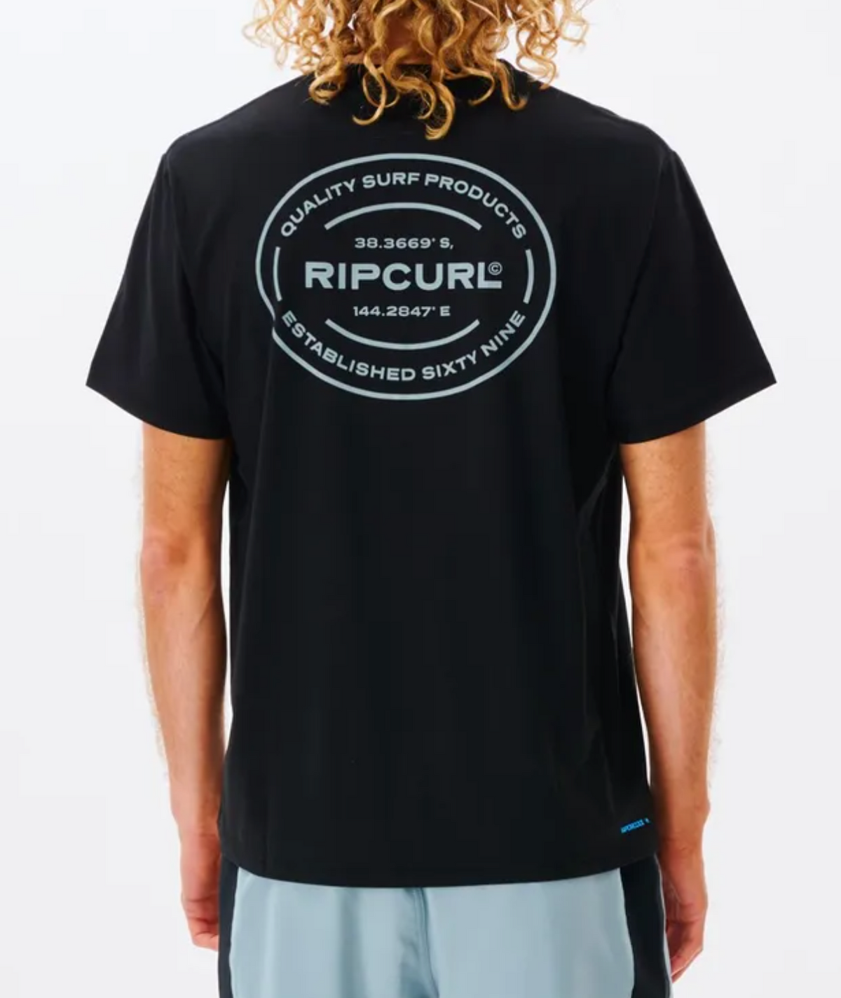 A person with curly blond hair wears a black Rip Curl Vaporcool Oval Tee featuring quick-dry fabric and a large circular back logo reading "Quality Surf Products," geographic coordinates, and "Established Sixty Nine.