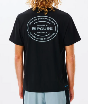 A person with curly blond hair wears a black Rip Curl Vaporcool Oval Tee featuring quick-dry fabric and a large circular back logo reading "Quality Surf Products," geographic coordinates, and "Established Sixty Nine.