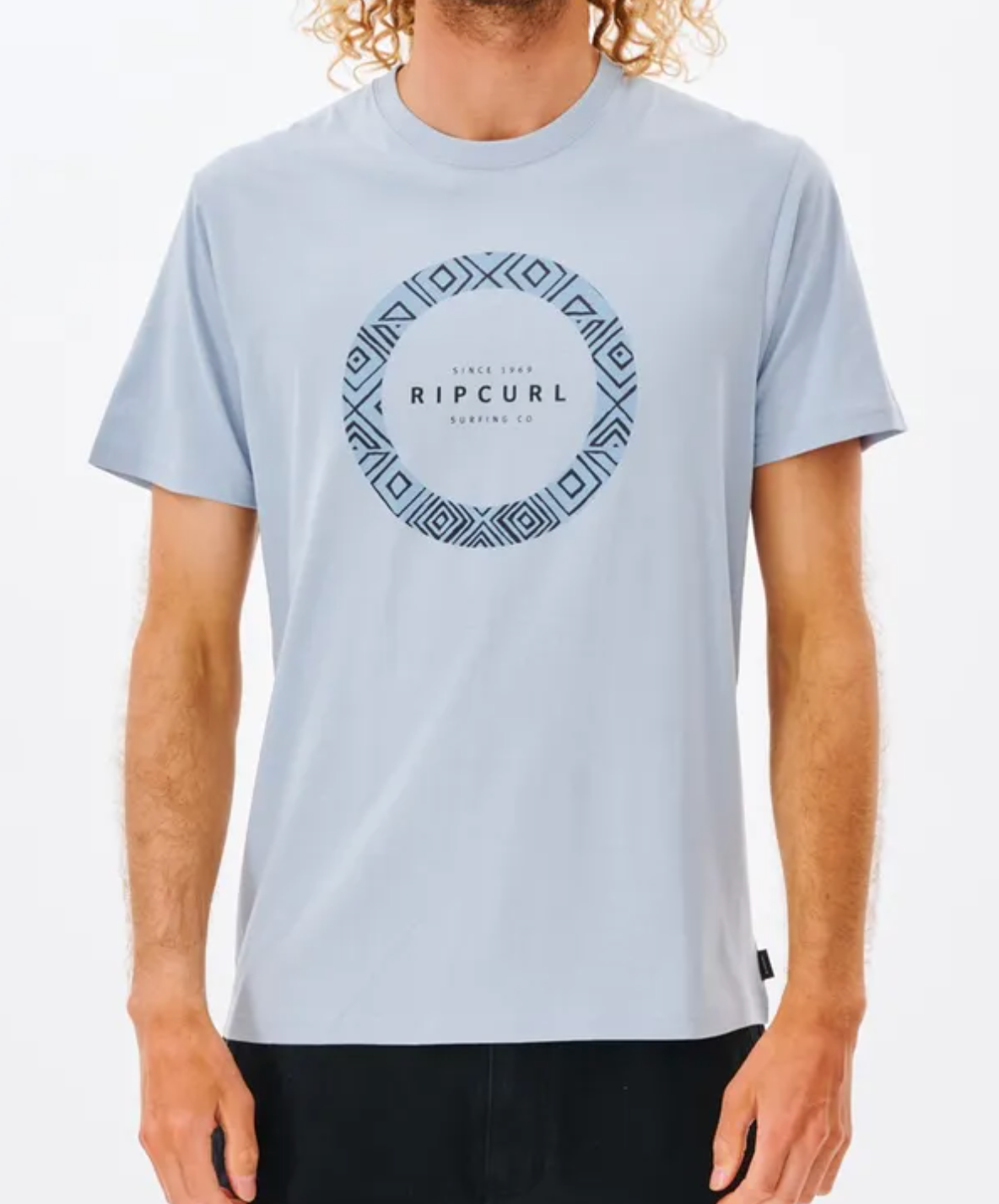 A person models a Rip Curl Fill Me Up Tee in light blue with a geometric pattern and logo on the front. The casual tee, made of 100% cotton, has short sleeves. The individual stands against a plain white backdrop.