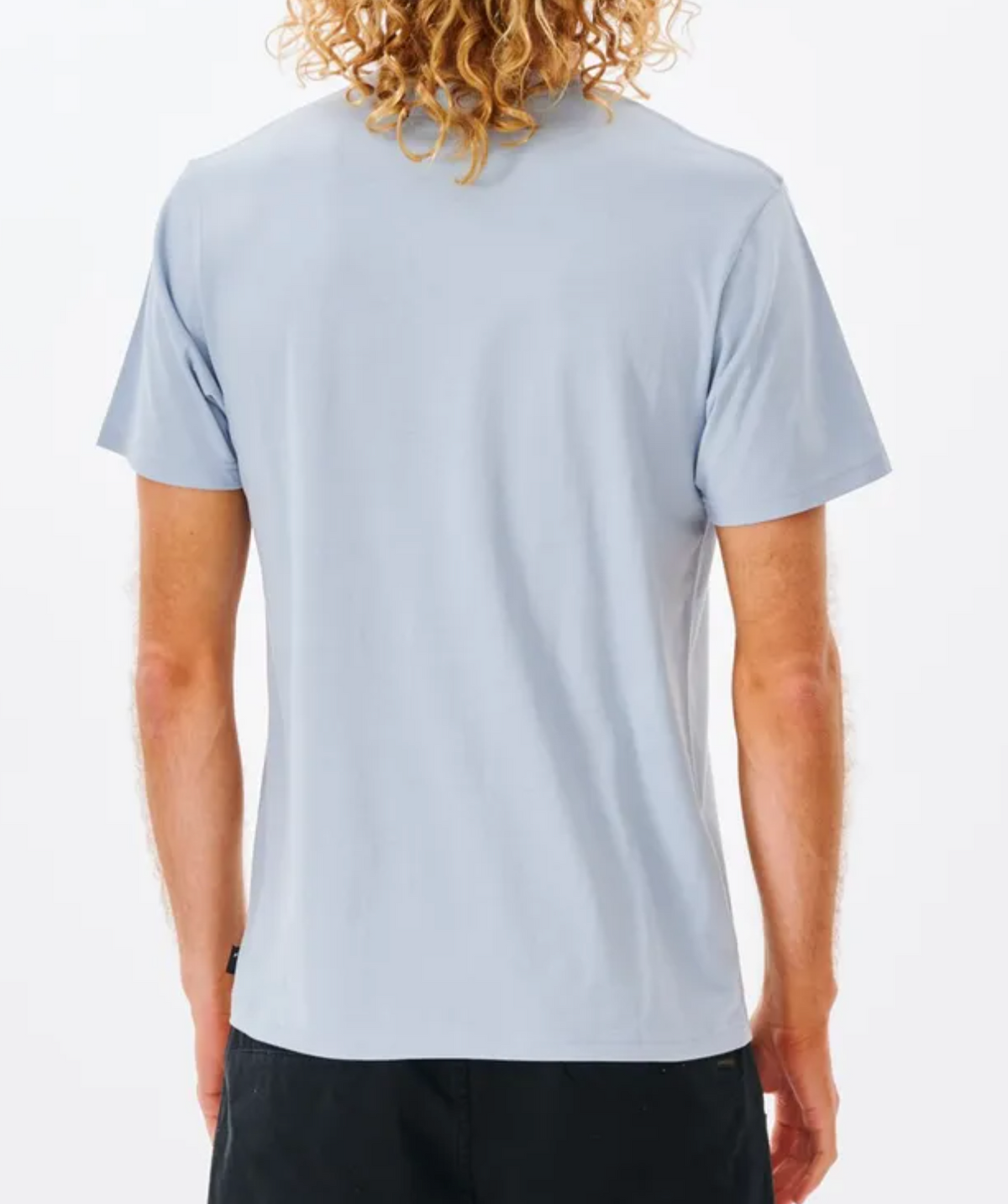 Rear view of a person with long curly hair wearing a light blue Rip Curl Fill Me Up Tee and black pants against a plain white background.