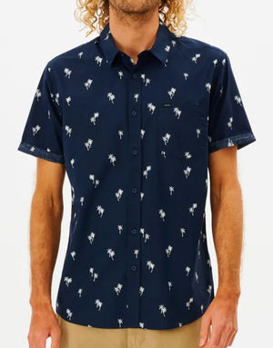 Wearing a navy Rip Curl Paradise Palms Shirt with palm tree patterns, short sleeves, button-up front, and chest pocket, a person stands against a plain backdrop. The shirt is 100% cotton and paired with light-colored pants.