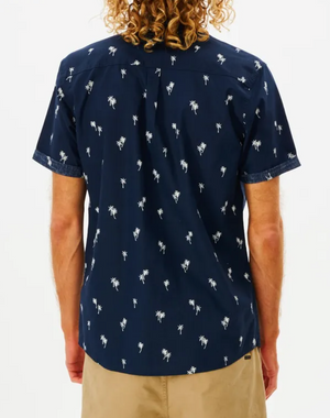 The rear view shows a curly-haired person in a navy blue Rip Curl Paradise Palms Shirt with short sleeves and a palm tree pattern, paired with khaki shorts. The 100% cotton ensemble stands out against a plain white background.