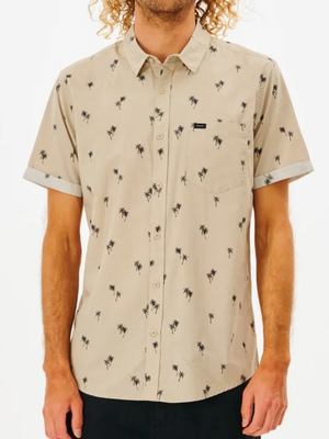 A person stands against a white background, wearing a Rip Curl Paradise Palms Shirt—a beige, short-sleeved, 100% cotton, button-up with a palm tree pattern. It features a front pocket and light sleeve trimming. Their curly hair hides their face.