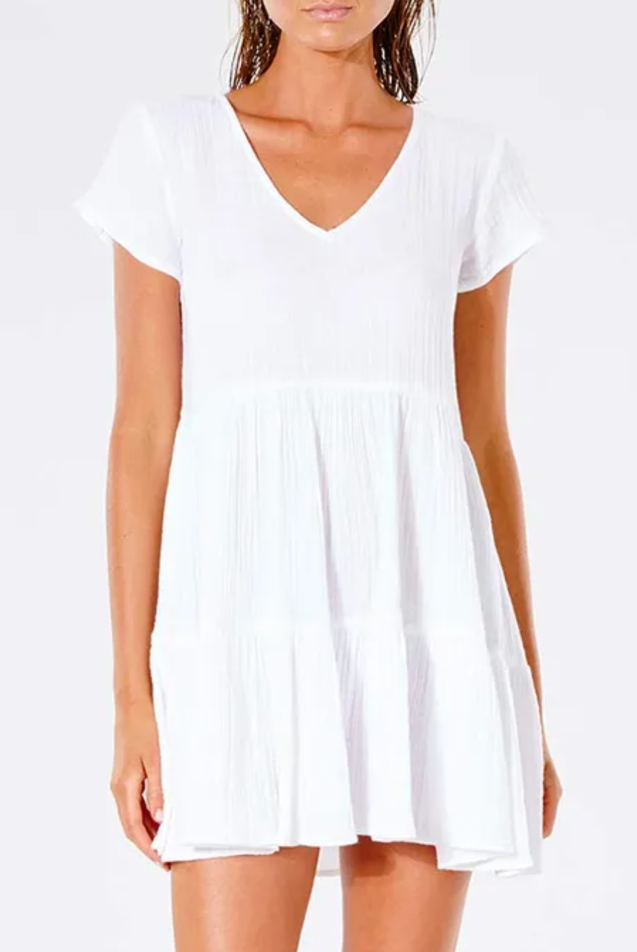 Someone wears a Rip Curl Premium Surf Dress, a white short-sleeved V-neck design, against a white background. This dress boasts a soft textured fabric with a loose, flowing skirt, ideal for casual occasions.