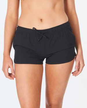 A person wears Rip Curl Classic Surf 3 Boardshort in black, crafted from recycled plastic bottles, against a white background. The focus is on the shorts with an elastic waistband and drawstring, made from 4-way stretch fabric. Visible is their lower torso to mid-thigh.