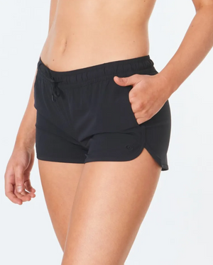 A person in Rip Curl Classic Surf 3 boardshorts, made from recycled bottles with 4-way stretch fabric, stands against a light background. The black shorts have an adjustable drawstring and a small logo near the hem, providing comfort.