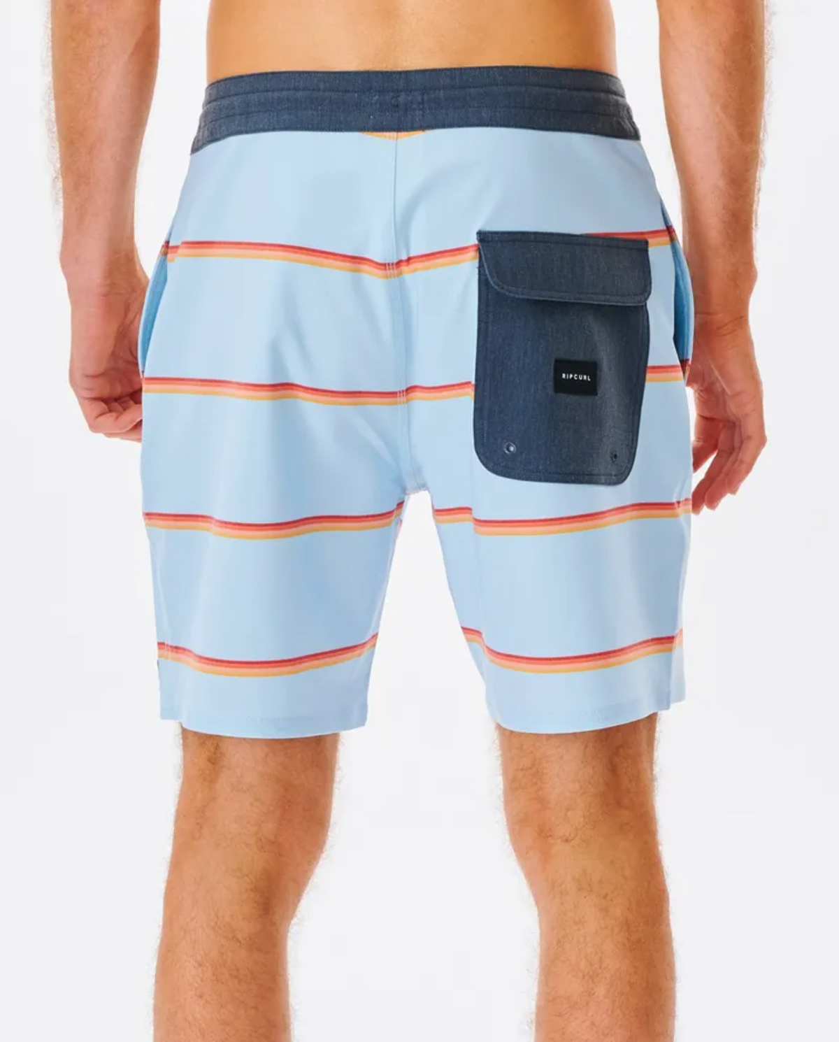 From behind, a person wears Rip Curl Monarch Layday boardshorts in light blue with red and yellow stripes, a dark blue waistband, and knee-length cut. Made from quick-drying recycled PET fabric, these shorts feature a large dark blue pocket with a small label for style.