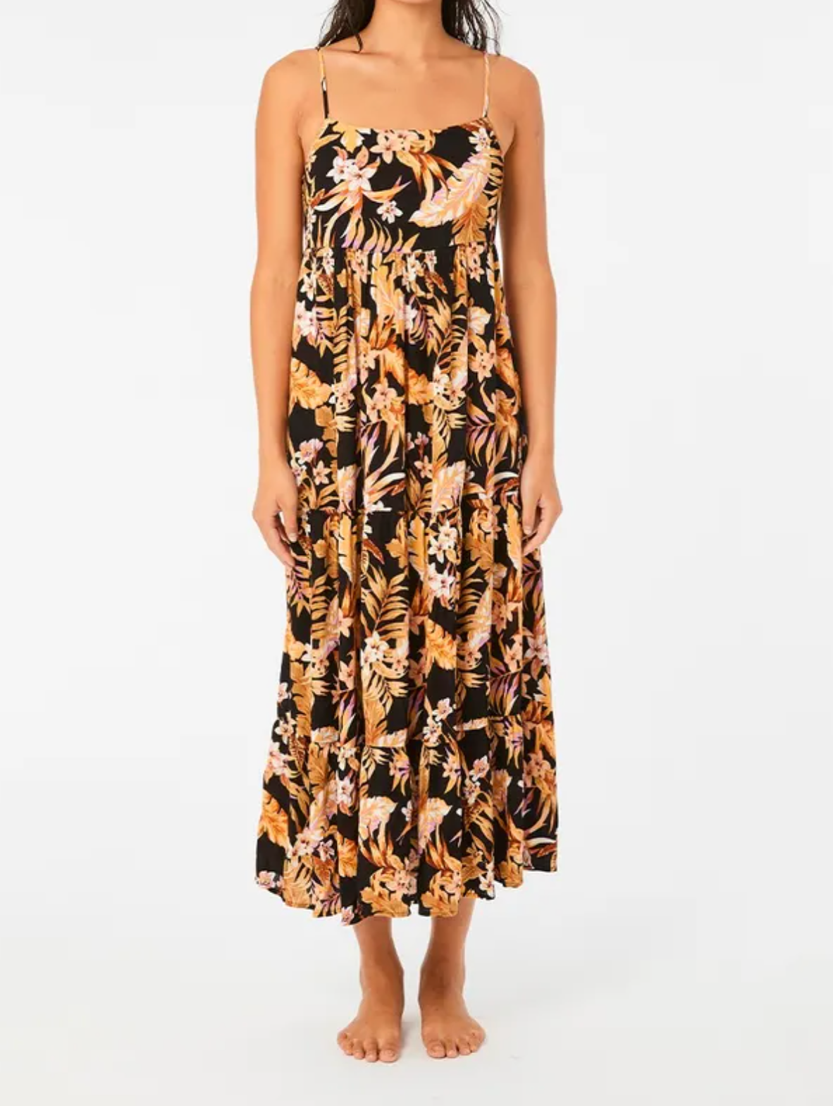 A person wearing the Rip Curl Sunday Swella Midi Dress, adorned with pink and orange flowers on a black backdrop, made from EcoVero fabric. This sustainable fashion piece has thin straps and flows to the ankles. The individual stands barefoot against a plain white backdrop.