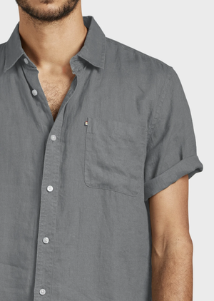 A person wears the Academy Brand Hampton Linen Short Sleeve Shirt in light gray, featuring a chest pocket and rolled sleeves. The comfortable shirt is slightly unbuttoned at the top, showing only the upper body and left arm.