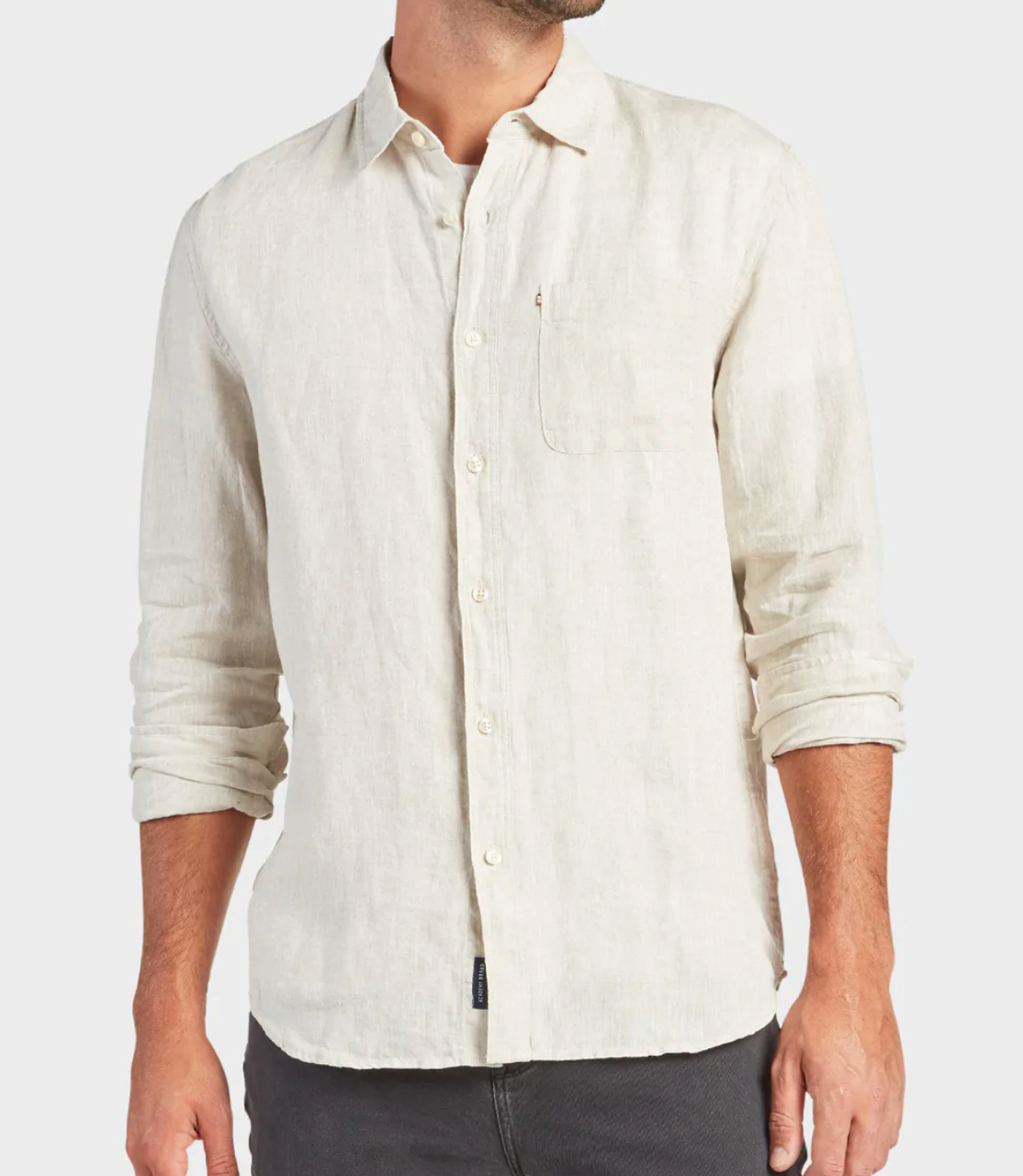 A man wears a regular-fit, beige Academy Brand Hampton Linen Shirt with a collar and chest pocket, sleeves slightly rolled up, paired with dark pants against a plain white background.