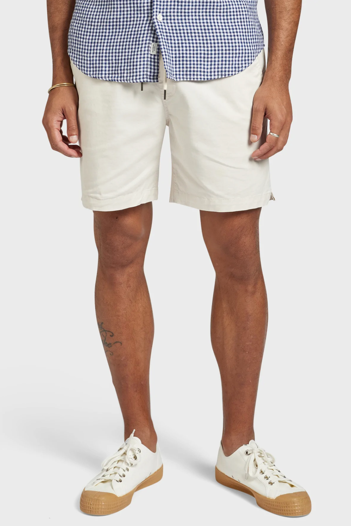 Sporting a blue and white checkered shirt, the person pairs it with beige Academy Brand Volley Shorts featuring an elastic drawstring waist and white sneakers. Their arms hang naturally, revealing a partially visible leg tattoo against a plain white background.