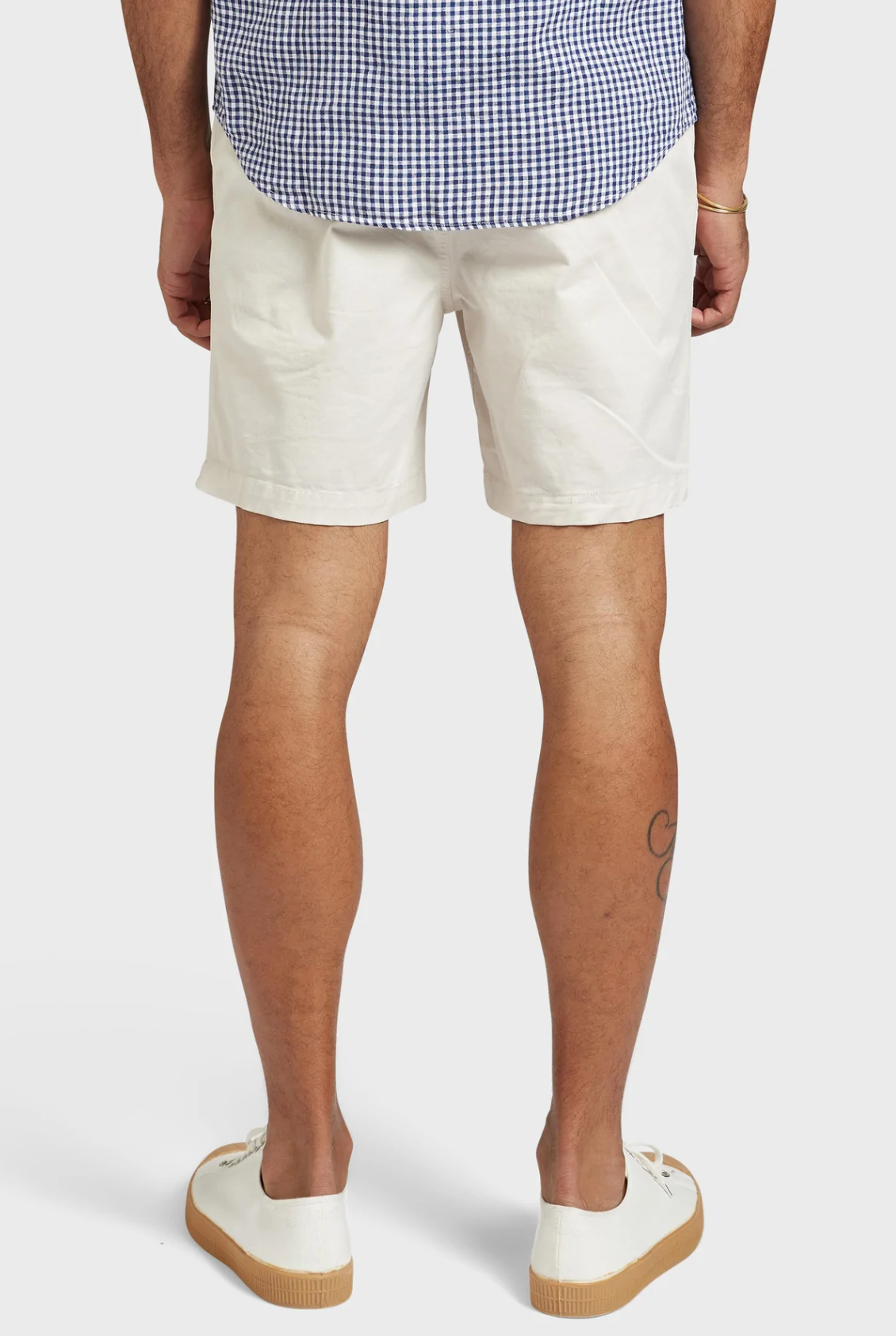 A person wearing a blue and white checkered shirt is seen from the rear, paired with Academy Brand Volley Shorts and white sneakers featuring brown soles. They have a tattoo on their right calf, set against a plain white background.