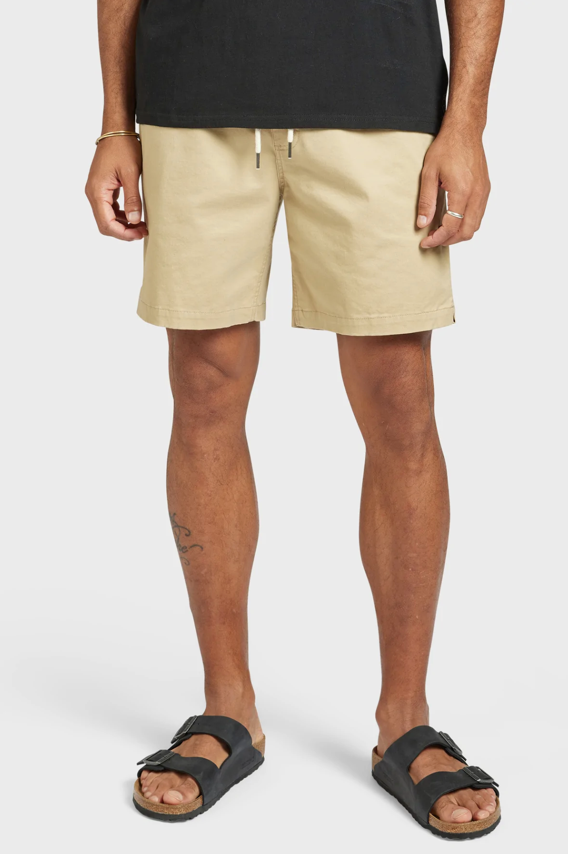 Against a white backdrop, a person sports the Academy Brand Volley Short in beige, featuring an elastic waist and white drawstrings, paired seamlessly with black slide sandals. They wear a black shirt, while their arms rest comfortably at their sides.