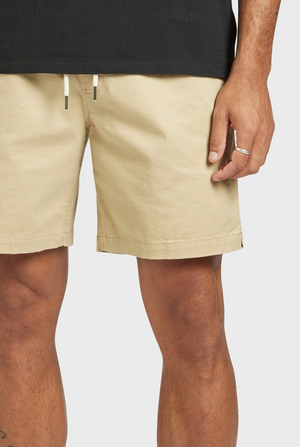 A person wears Academy Brand Volley Shorts in beige, featuring an elastic waist and drawstring, paired with a black shirt. A silver ring adorns their hand by their side against a plain white background.