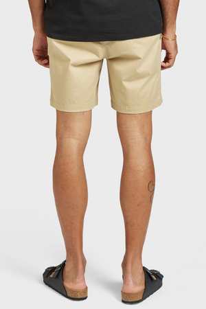 A person stands with their back to the camera wearing a black shirt, Academy Brand Volley Short with an elastic drawstring waist, and black sandals. A small tattoo is visible on the back of their right calf.