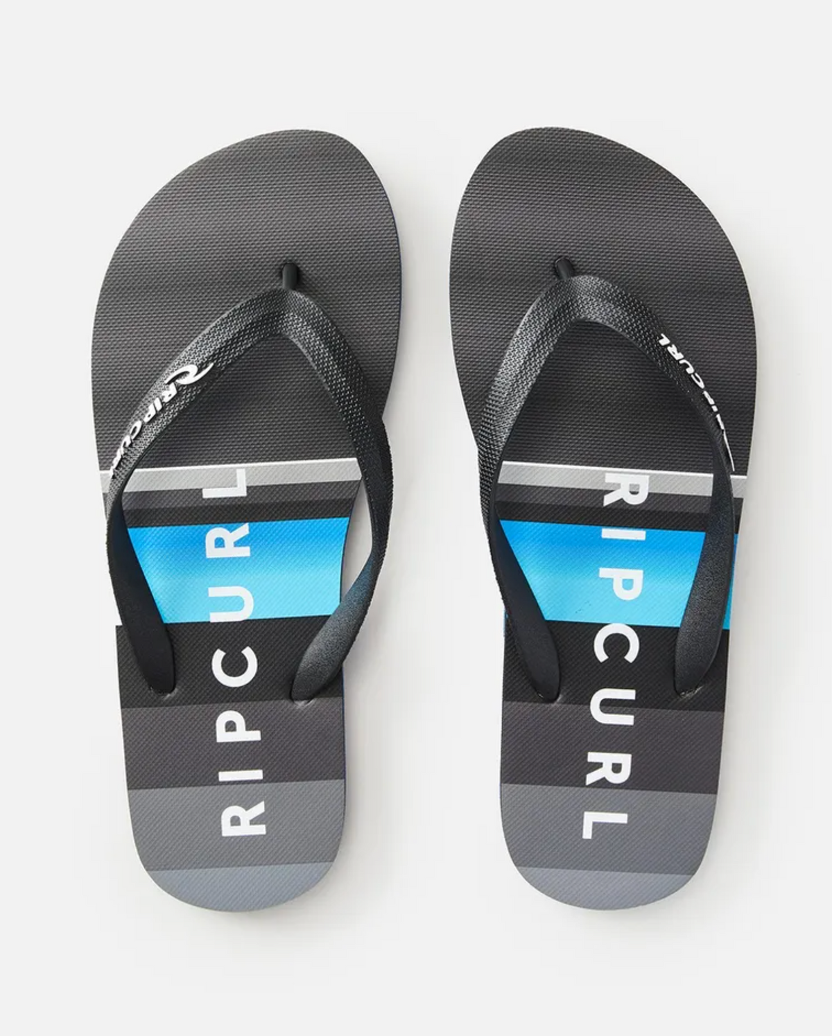 The Rip Curl Daybreak Thongs boast an ergonomic molded rubber strap for comfort, with soles made from premium EVA rubber featuring horizontal gray, blue, and black stripes with "RIP CURL" boldly printed in white across the center.