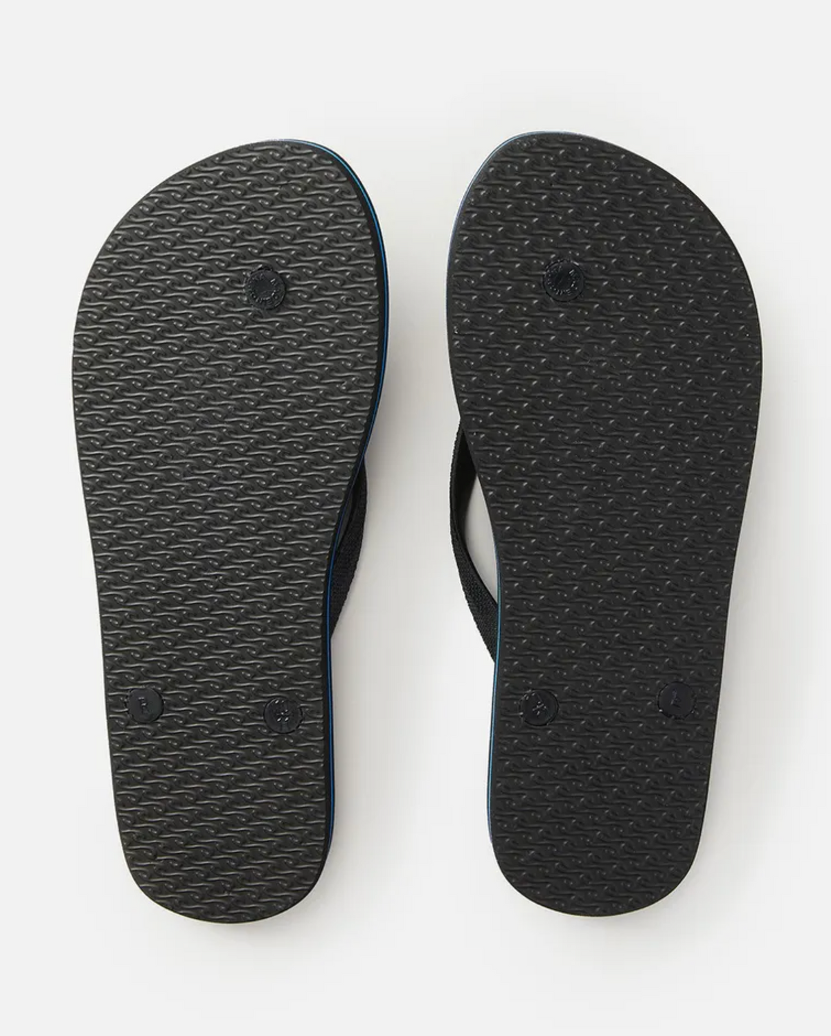 A pair of Rip Curl Daybreak Thongs photographed from the bottom, showcasing the tread pattern on the black soles. The textured soles feature a diagonal line design and circular impressions. These flip-flops have an ergonomic molded rubber strap and a premium EVA rubber footbed.
