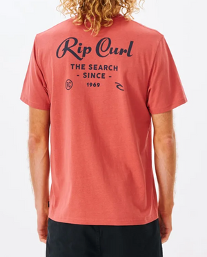 A curly-haired person wears a Rip Curl Twist Tee in red, featuring bold black "The Search Since 1969" text on the back, paired with black pants, standing confidently against a white background.
