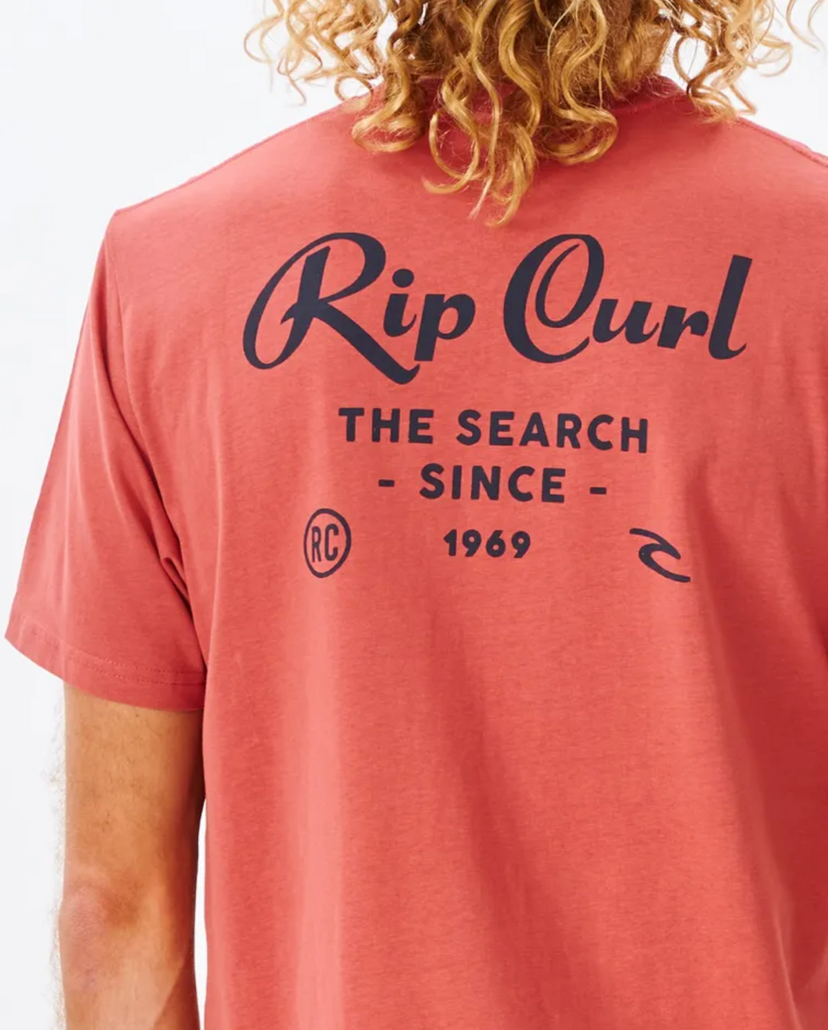 A person with curly hair is wearing a comfy red Rip Curl Twist Tee, featuring "Rip Curl" and "The Search Since 1969" printed in black on the back.