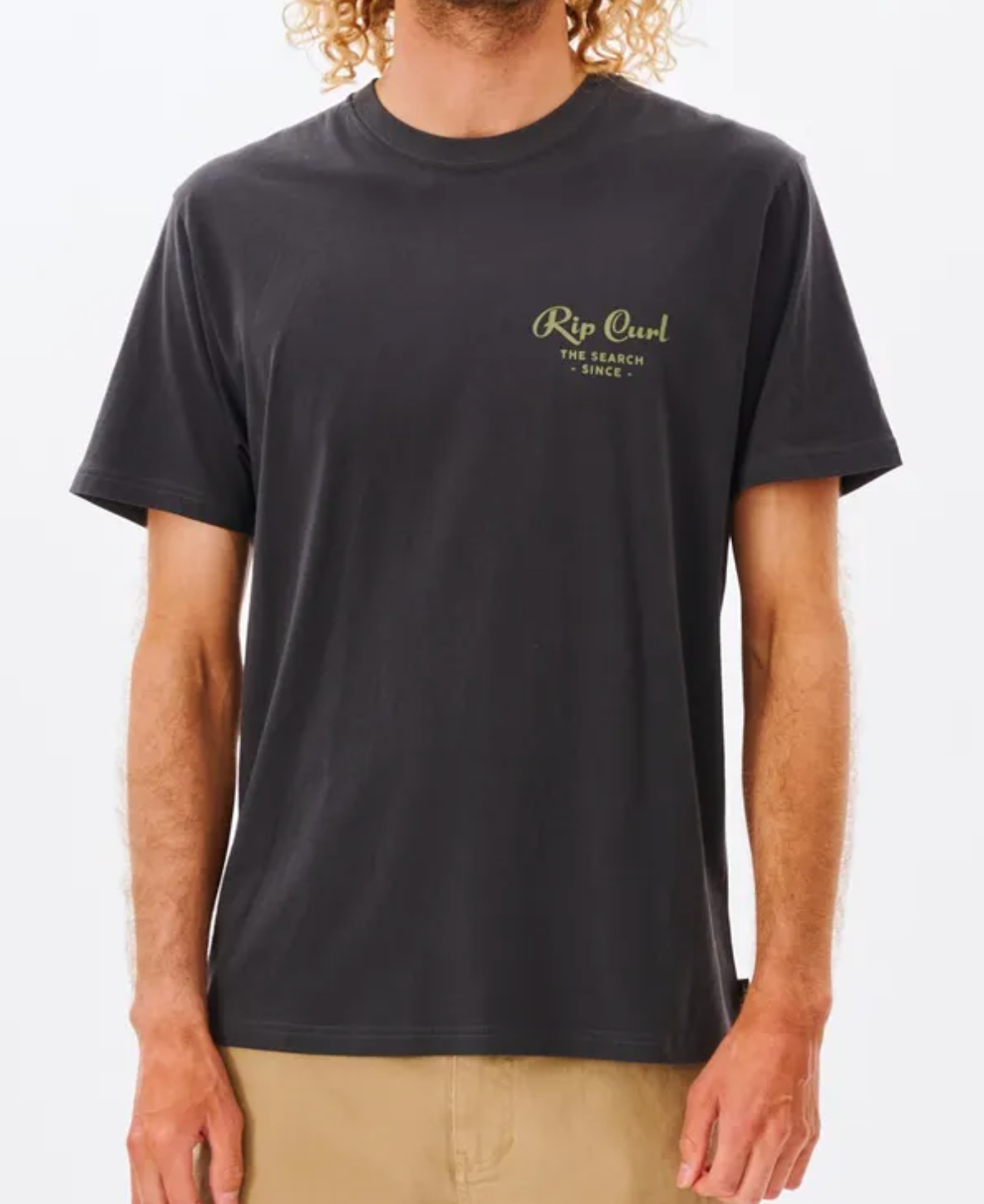 Wearing a comfy Rip Curl Twist Tee with "The Search Since..." in green on the left chest, a person complements their curly hair with beige pants.
