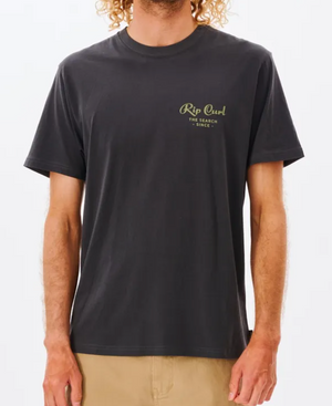 Wearing a comfy Rip Curl Twist Tee with "The Search Since..." in green on the left chest, a person complements their curly hair with beige pants.