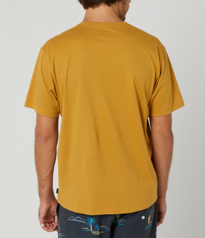 A man faces away from the camera, wearing a mustard yellow Rip Curl Corp Icon Tee made of 100% cotton and dark tropical print shorts. The background is plain and light-colored.