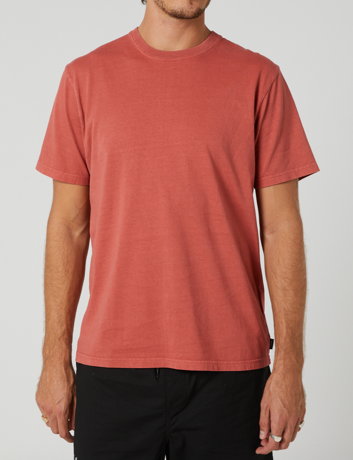 A person is wearing a Rip Curl Plain Wash Tee, a short-sleeved, 100% cotton faded red t-shirt, paired with black pants. The relaxed fit of the tee enhances comfort as the person stands against a plain light gray background. The photo is cropped at the shoulders and upper thighs.
