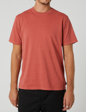 A person is wearing a Rip Curl Plain Wash Tee, a short-sleeved, 100% cotton faded red t-shirt, paired with black pants. The relaxed fit of the tee enhances comfort as the person stands against a plain light gray background. The photo is cropped at the shoulders and upper thighs.