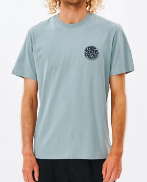A person with curly hair wearing the Rip Curl Wetsuit Icon Tee in a light blue short-sleeve style, featuring a round black Rip Curl print on the left chest, stands against a plain white background. This comfortable 100% cotton tee pairs perfectly with dark pants.