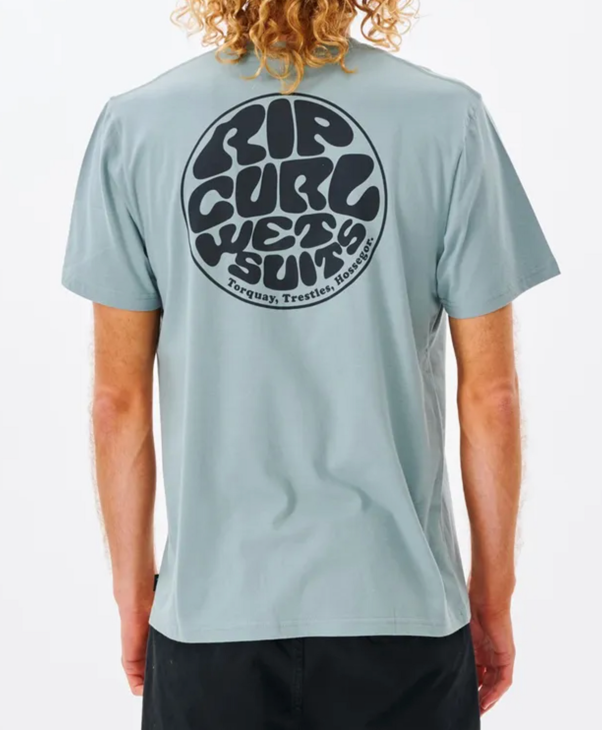 A person with curly blonde hair is wearing the Rip Curl Wetsuit Icon Tee, a light blue T-shirt made of 100% cotton. The shirt features a large print graphic on the back with the words "RIP CURL WET SUITS" and location names "Torquay, Trestles, Hossegor" in bold, retro style font.