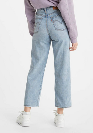 Facing away from the camera, a person wears high-rise Levi's Ribcage Straight Jeans in light blue with a lavender sweatshirt. The slim-fit jeans feature a visible leather patch on the waistband and are paired with white sneakers against a plain white background.