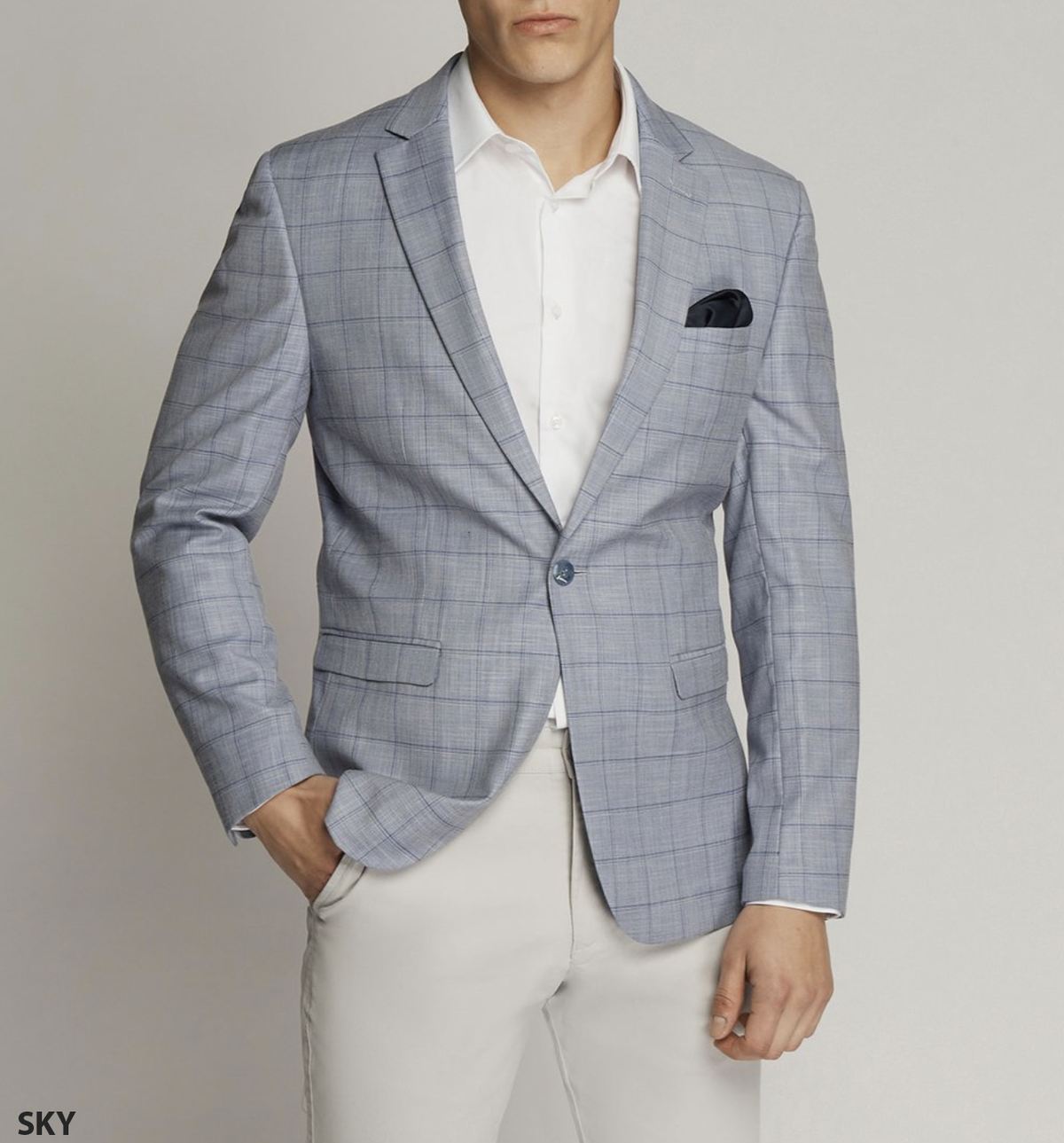 A person in a Scuzzatti Check Slim Fit Sports Jacket, light gray with a checkered pattern, paired with a white shirt and beige pants. A black pocket square is neatly tucked into the blazer's breast pocket, showcasing precise styling against the plain light gray backdrop.