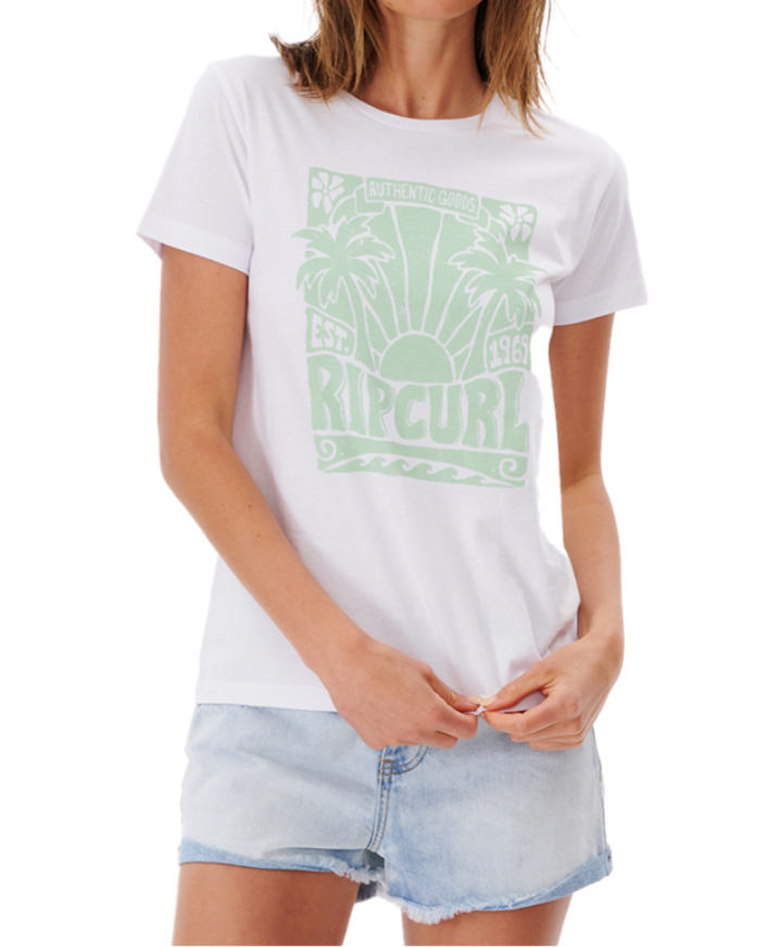 Wearing a Rip Curl Sol Seeker Standard Tee in organic cotton with a light green palm tree design and "RIP CURL, EST. 1969," the person pairs it beautifully with light denim shorts against a plain backdrop.