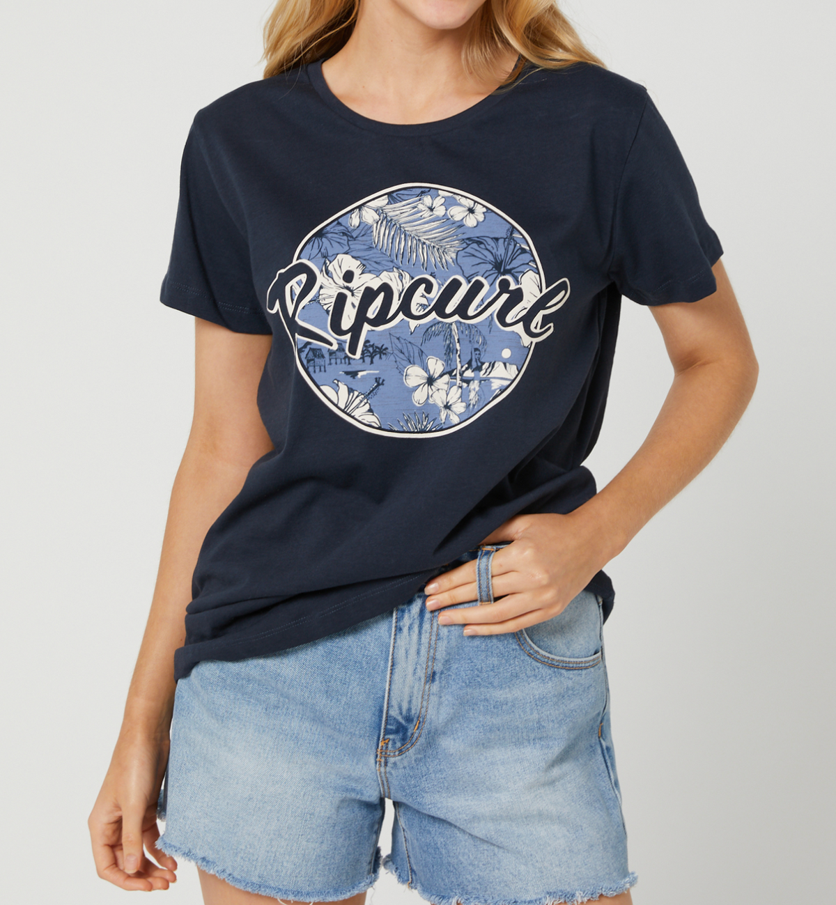 Someone wearing a Rip Curl Beachside Standard Tee in navy blue, made from 100% cotton with a floral graphic, paired with denim shorts, stands against a plain light background.
