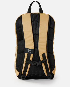 The Rip Curl Overtime 30L Overland Backpack in tan and black has padded shoulder straps, a chest buckle, breathable black mesh back, made from recycled polyester. It supports 15" laptops, features adjustable carrying straps, and includes a small logo patch on one strap.