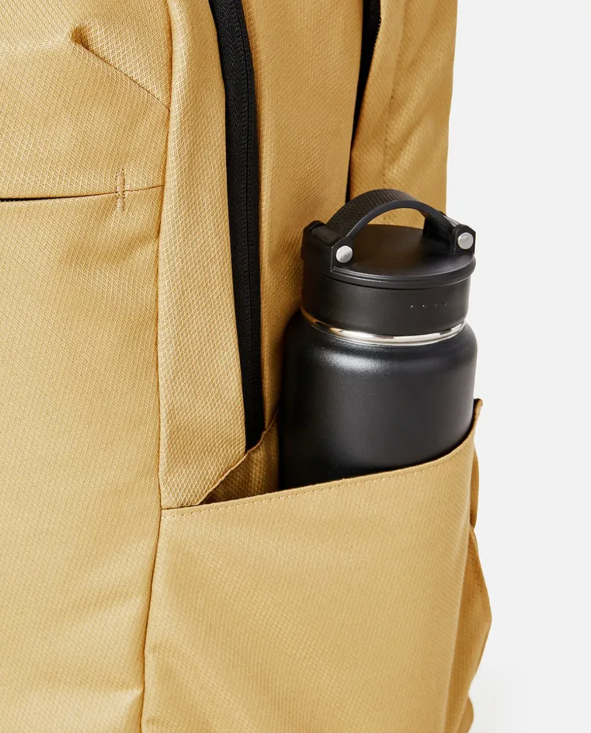 A black water bottle with a handle is partially tucked into the side pocket of a Rip Curl Overtime 30L Overland Backpack, made from recycled polyester and designed to fit a 15" laptop.
